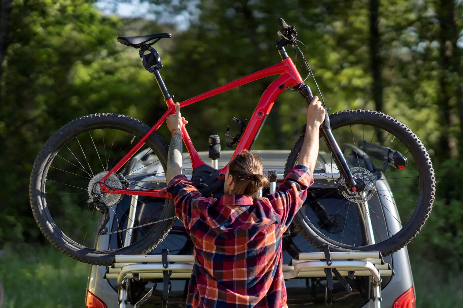 Rückansicht eines Mannes mit zum Dutt gebundenen Haaren in einem rot-blau karierten Flanellhemd, der ein leuchtend rotes Hardtail-Mountainbike auf einen silbernen Heckklappenträger eines grauen Vans hebt. Das Fahrrad befindet sich quer zur Fahrtrichtung, im Hintergrund ist ein dicht bewachsener, grüner Wald in hellem Tageslicht zu sehen.