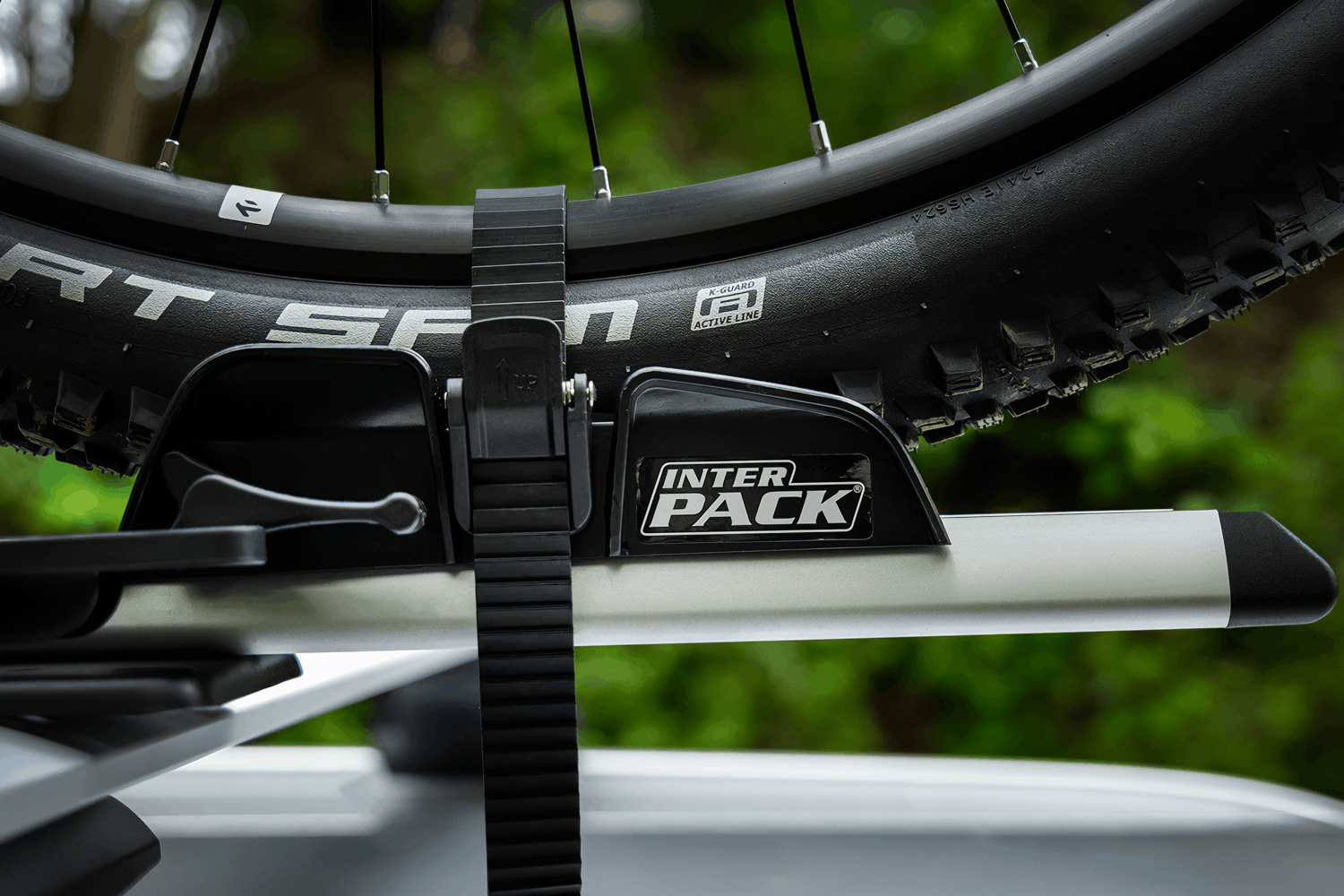 Detaillierte Nahaufnahme eines Mountainbike-Reifens der Marke Schwalbe (Modell Smart Sam), der in der Kunststoff-Radschale eines Fahrradträgers steht. Ein schwarzes Kunststoff-Ratschenband ist fest um die Felge gezogen. Auf der Halterung prangt gut sichtbar das weiße Logo von Inter Pack auf schwarzem Grund. Die Trägerschiene besteht aus mattem Aluminium mit einer schwarzen Endkappe, der Hintergrund ist unscharf grün.