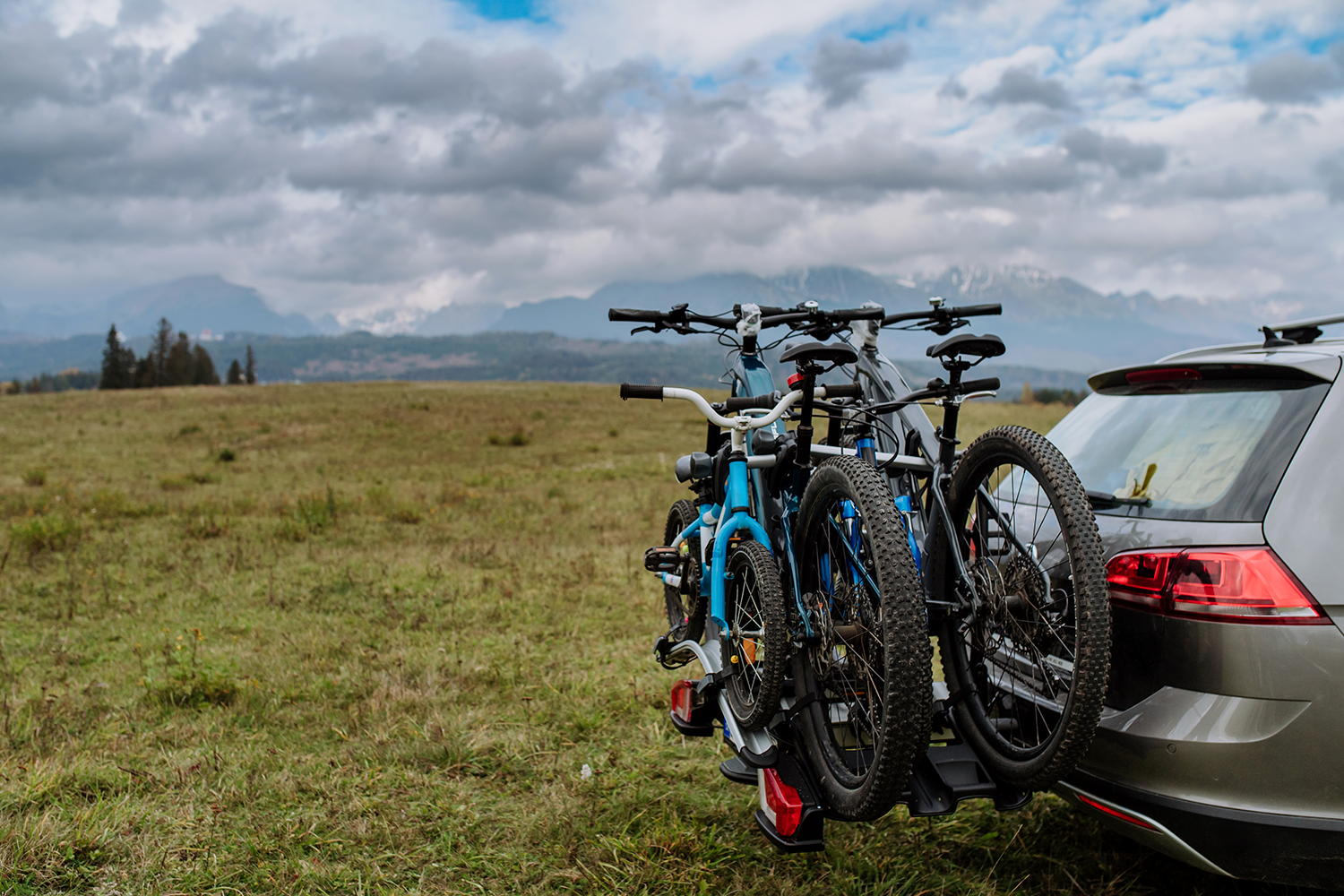 Drei Fahrräder auf einem Fahrradträger. Der Träger ist an der Anhängerkupplung eines beigen Autos befestigt. Im Hintergrund eine bergige, grüne Landschaft und ein Himmel voller weiß-grauer Wol