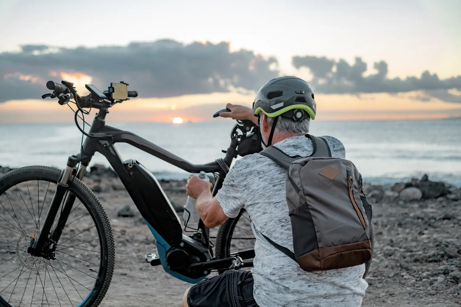 Rückansicht eines aktiven, älteren Mannes mit grauem Haar, der mit einem Fahrradhelm und einem grau-braunen Rucksack an einer felsigen Meeresküste neben seinem E-Mountainbike hockt. Er bedient eine Trinkflasche am Rahmen des dunklen Elektrofahrrads, das mit einer Smartphone-Halterung am Lenker ausgestattet ist. Im Hintergrund spiegelt sich ein stimmungsvoller Sonnenuntergang über dem ruhigen Meer, während die Sonne tief am Horizont unter einer dichten Wolkendecke hervor bricht.