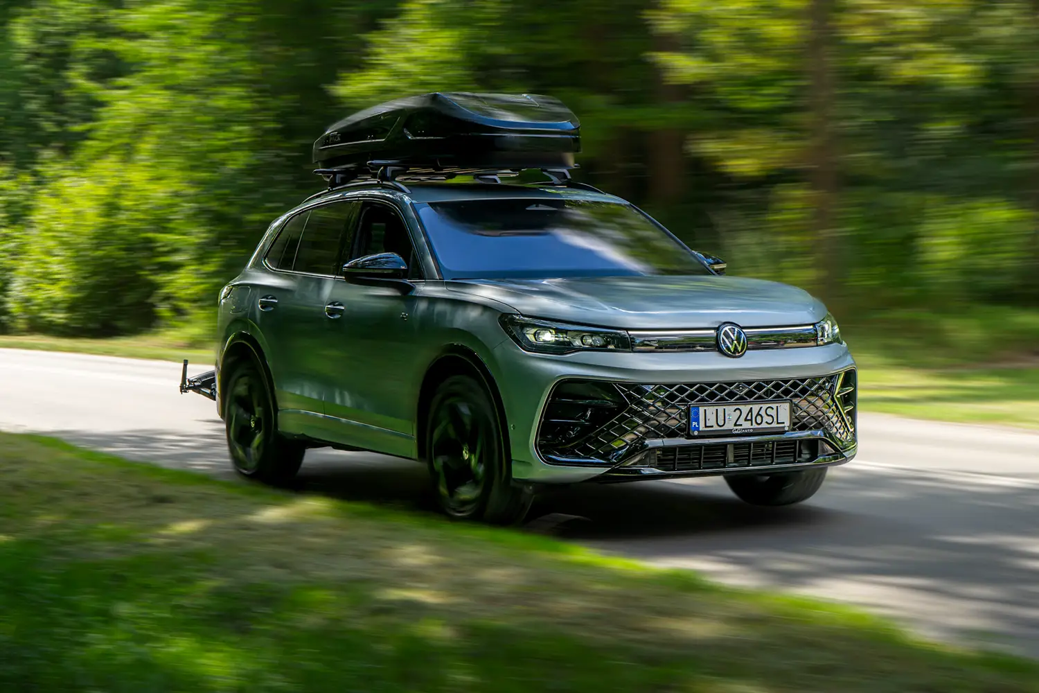Ein silberner SUV mit Volkswagen-Logo, von vorne gesehen. Das Auto ist in Bewegung. Auf dem Dach ist eine schwarze Dachbox montiert, und an der Anhängerkupplung befindet sich ein Fahrradträger. Eine schmale Asphaltstraße, zu beiden Seiten ist Wald zu sehen.