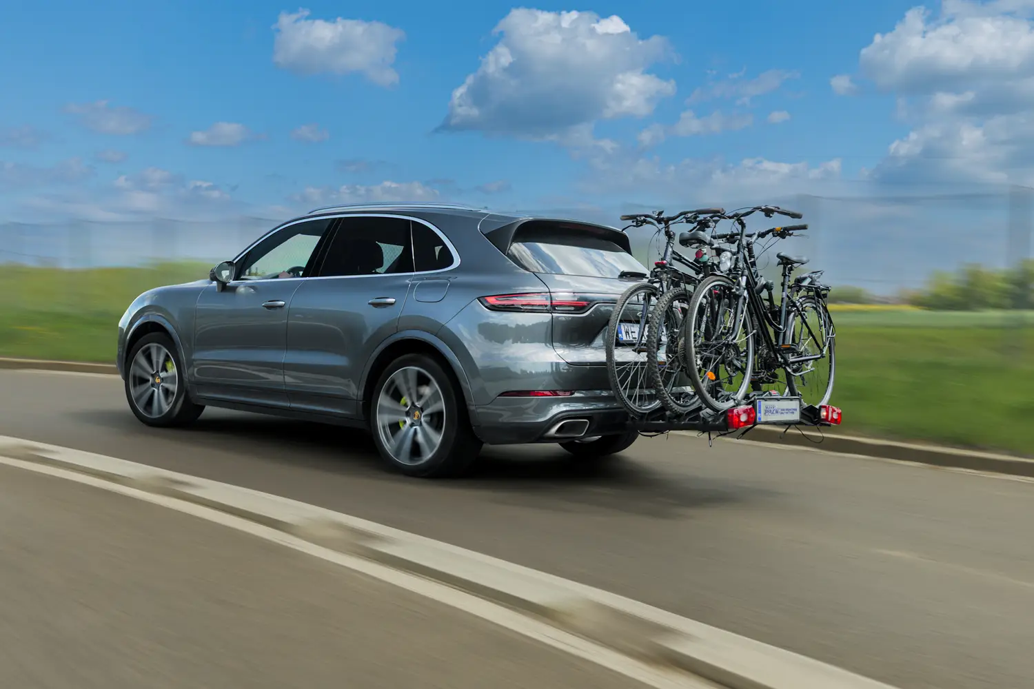 Ein silberner SUV mit montierter Fahrradträger. Auf dem Träger sind drei schwarze Fahrräder befestigt. Das Auto fährt auf einer asphaltierten Straße. Im Hintergrund sind Grünflächen und ein klarer Himmel zu sehen.