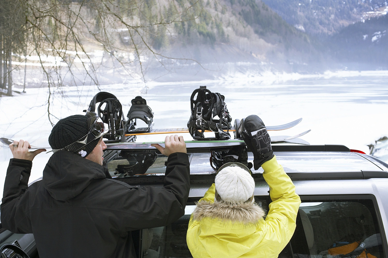 Eine Frau und ein Mann laden Snowboards von einem Dachgepäckträger. Beide tragen Winterjacken und Mützen. Der Mann ist schwarz, die Frau gelb gekleidet. Im Hintergrund erstreckt sich eine Winterlandschaft mit See, Wald und Hügeln.