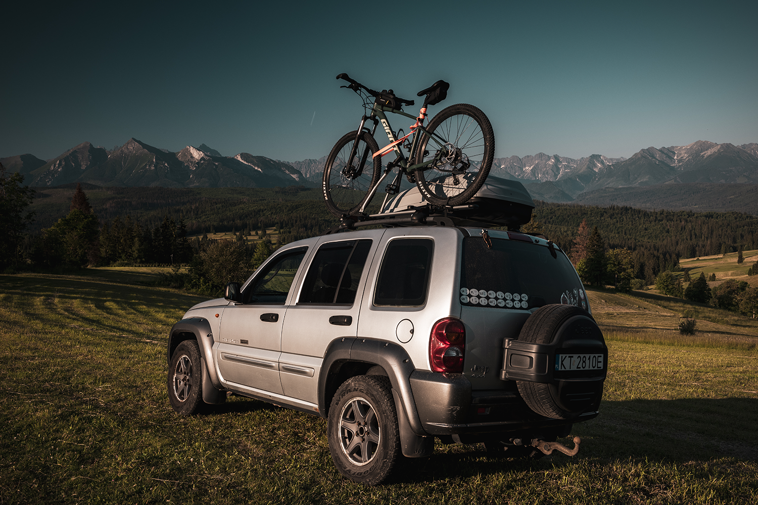  Ein silberner Geländewagen auf einem Feld, im Hintergrund Berge und ein Wald. Auf dem Dach des Wagens sind ein Fahrrad und eine Dachbox montiert