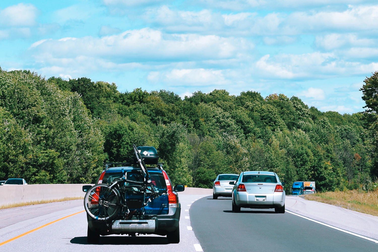 Ein schwarzes Auto fährt auf der linken Spur der Autobahn. Auf der linken Spur befinden sich zwei helle Autos. Auf der Heckklappe des schwarzen Autos ist ein Fahrrad befestigt, und auf dem Dach befindet sich eine Dachbox. Auf beiden Seiten der Straße erstreckt sich ein Wald. Es ist ein sonniger Tag.