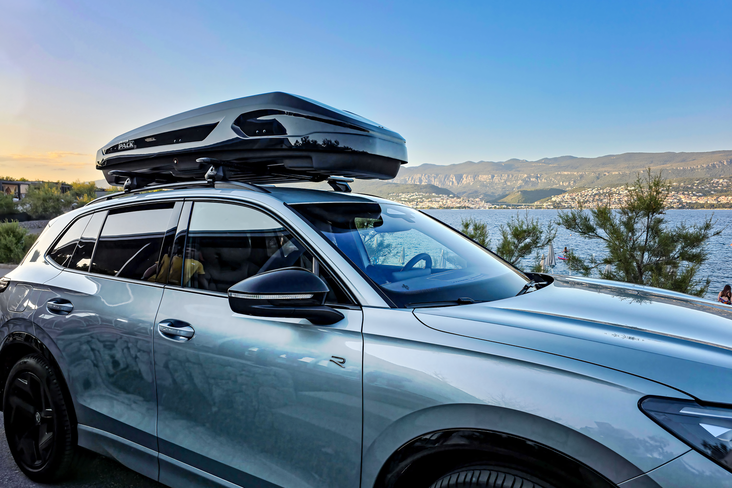Nahaufnahme von rechts auf ein silbernes Auto mit einer montierten schwarzen Dachbox. Das Auto und die Box sind mit einer Hochglanzlackierung versehen. Im Hintergrund ist die Landschaft einer Küstenstadt zu sehen. Der Himmel ist klar und weist Blau- und Orangetöne auf.