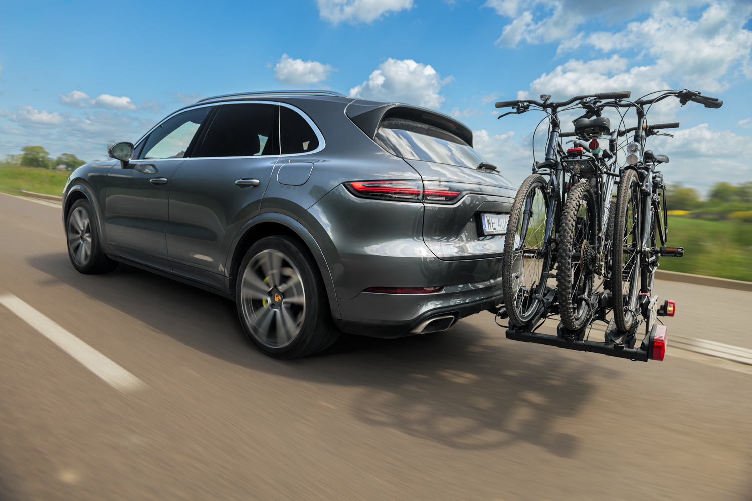  Ein silberner SUV, der auf einer zweispurigen Straße fährt und einen Fahrradträger an der Anhängerkupplung hat. Auf der Ladefläche sind drei Mountainbikes befestigt.