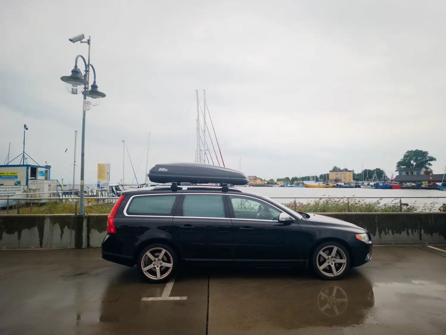 Ein schwarzer Kombi mit einer montierten Dachbox parkt an einer Uferpromenade in der Nähe eines Hafens mit Segelbooten unter bedecktem Himmel