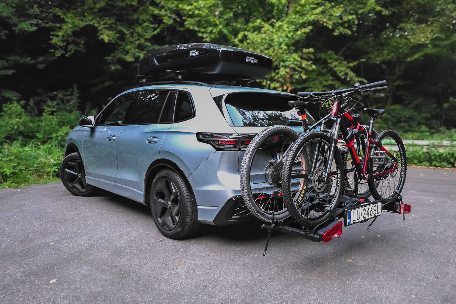 Ein moderner, hellblauer SUV parkt auf einer grauen Asphaltstraße vor einem dichten, grünen Wald. Auf dem Fahrzeugdach ist eine glänzende, aerodynamische schwarze Dachbox der Marke Inter Pack montiert. Am Heck befindet sich ein schwarzer Anhängerkupplungsträger, auf dem zwei Mountainbikes – ein auffälliges rot-schwarzes und ein dunkles – nebeneinander befestigt sind. Das Kennzeichen „LU 246SL“ sowie die roten Rückleuchten des Trägers sind deutlich erkennbar.