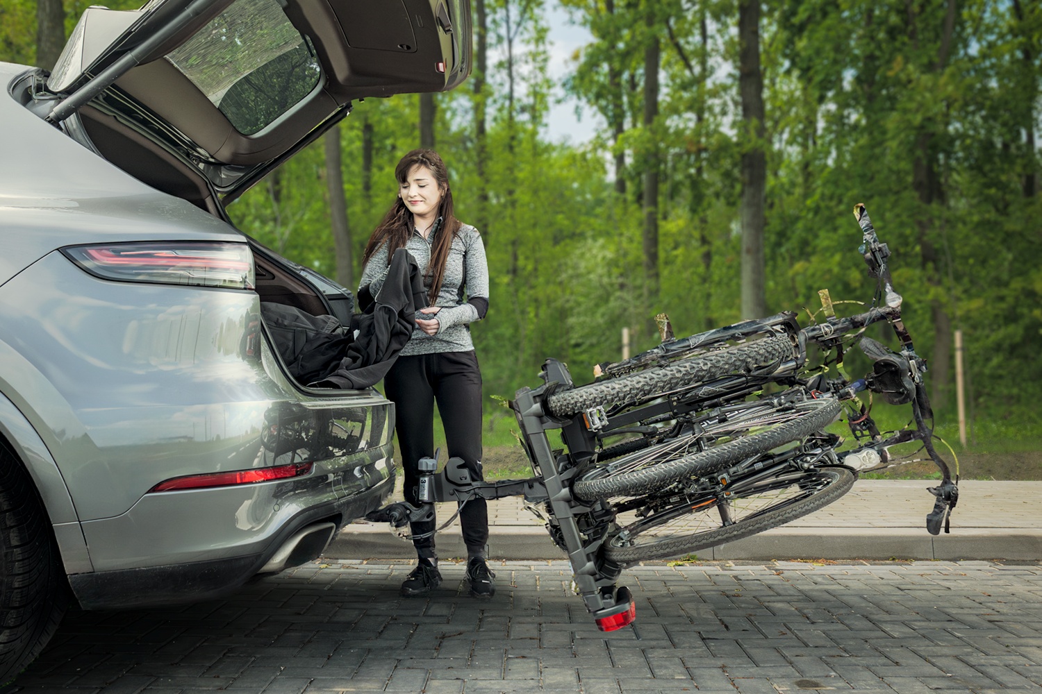 Eine junge Frau mit langen, dunklen Haaren in einer grauen Sportjacke und schwarzer Hose steht am Heck eines grauen SUVs. Die große Kofferraumklappe des Autos ist komplett nach oben geöffnet. Der auf der Anhängerkupplung montierte Fahrradträger, auf dem Fahrräder befestigt sind, ist weit nach hinten und unten weggeklappt, sodass er fast den gepflasterten Boden berührt und den Zugang zum Kofferraum freigibt. Im Hintergrund ist ein Wald mit hohen, grünen Bäumen zu sehen.