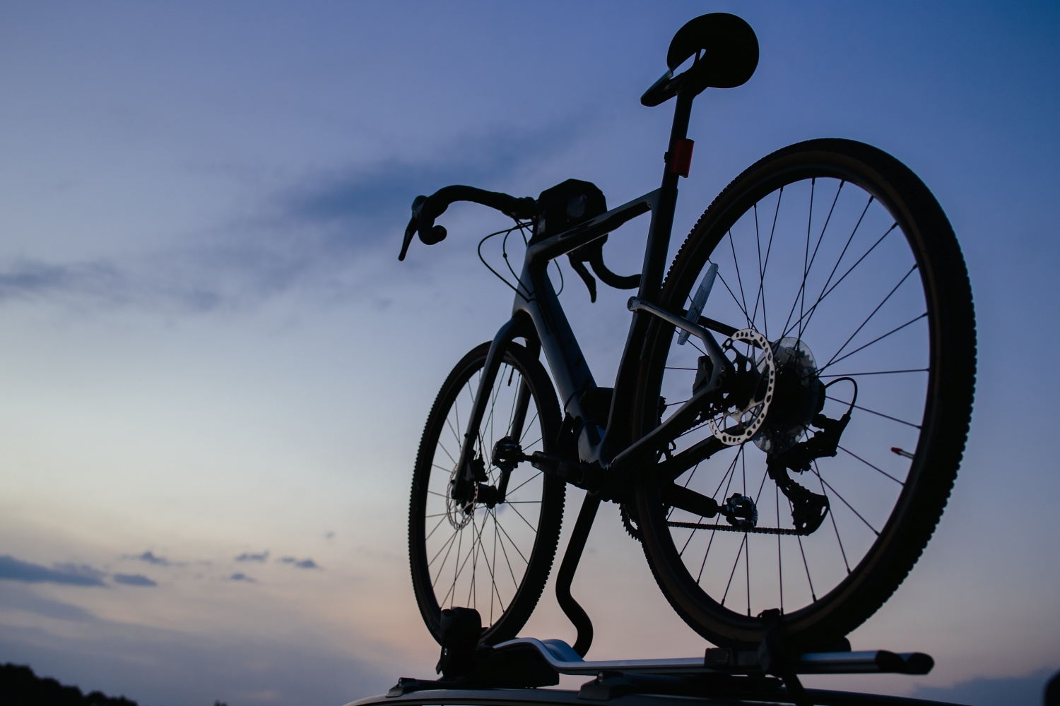 Eine detaillierte Silhouette eines sportlichen Gravel-Bikes mit geschwungenem Rennlenker, sichtbarer Gangschaltung und markanten Scheibenbremsen, aus einer niedrigen Perspektive von unten fotografiert. Das Fahrrad ist auf einem silbernen Dachträger fixiert und zeichnet sich scharf vor einem sanften, blau-violetten Abendhimmel im Zwielicht ab.
