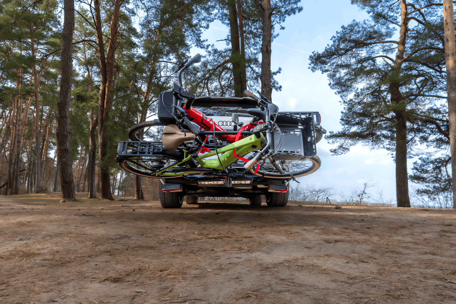 Ein Wald, im Hintergrund ein See. Im Mittelpunkt des Bildes steht ein silberner SUV mit einem Fahrradträger, der ausgeklappt ist. Auf dem Träger sind zwei E-Bikes in den Farben Rot und Grün befestigt.