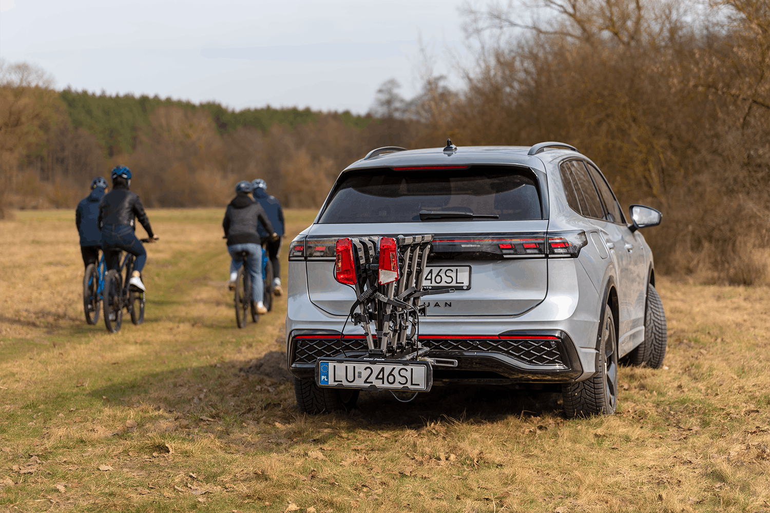 Eine Lichtung mitten im Wald, herbstliche Farben. Im Mittelpunkt des Bildes ein silberner SUV mit einem heruntergeklappten Fahrradträger. Im Hintergrund eine Gruppe von vier Personen auf Fahrrädern, die sich in die Ferne entfernt