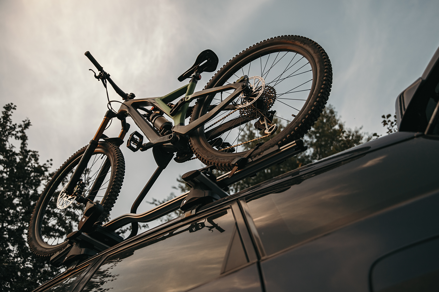 Dynamische Froschperspektive auf ein olivgrün-schwarzes Full-Suspension-Mountainbike, das auf einem Dachfahrradträger eines dunklen Autos montiert ist. Das Fahrrad mit seinen profilierten Reifen und der präzisen Schaltung hebt sich scharf von einem bewölkten Nachmittagshimmel ab.