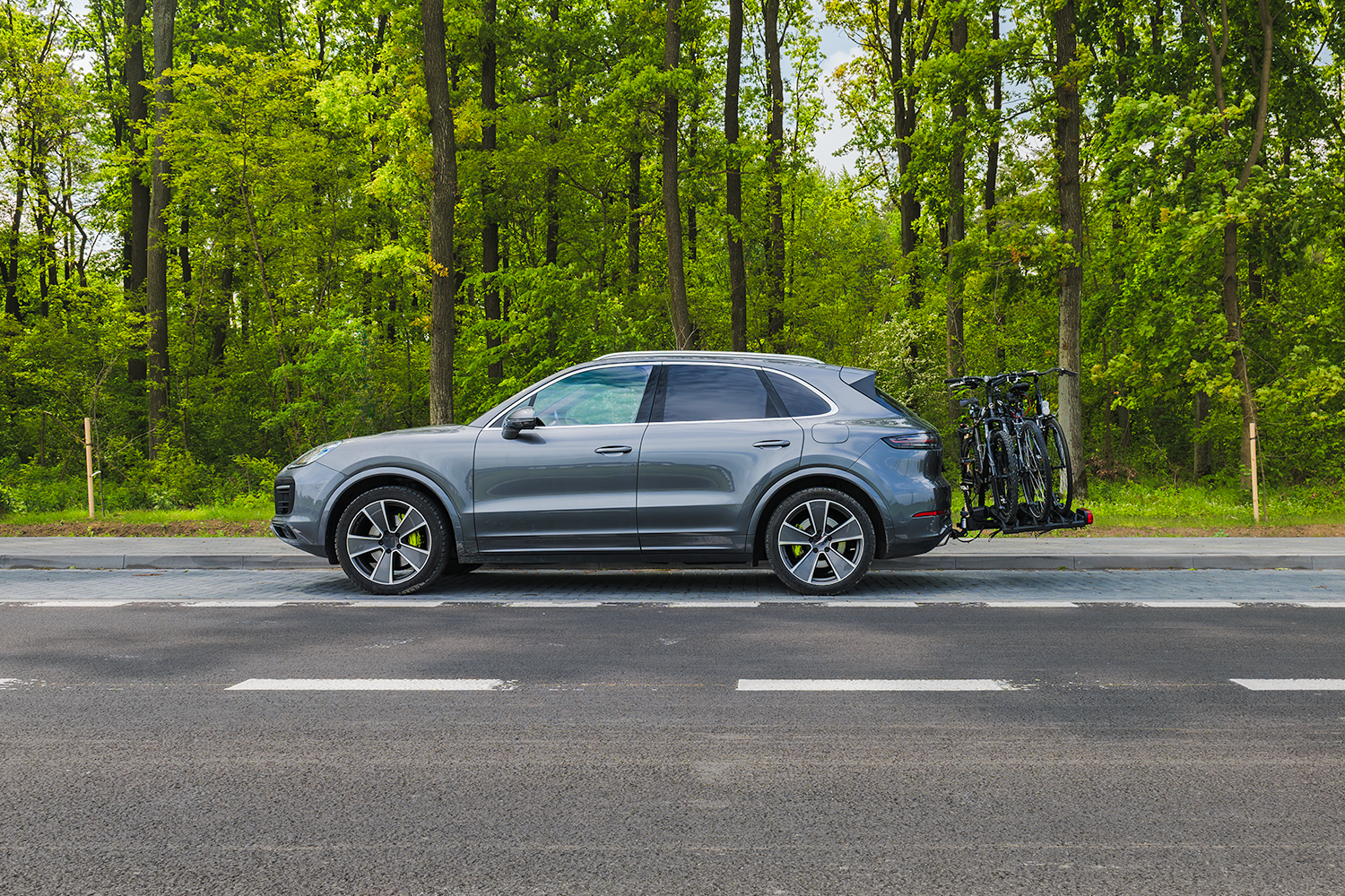 Seitenansicht eines grauen SUVs, der auf einem gepflasterten Streifen neben einer asphaltierten Straße vor einem dichten, grünen Wald steht. Am Heck des Fahrzeugs ist ein robuster Anhängerkupplungs-Fahrradträger montiert, auf dem drei schwarze Fahrräder für den Transport befestigt sind.
