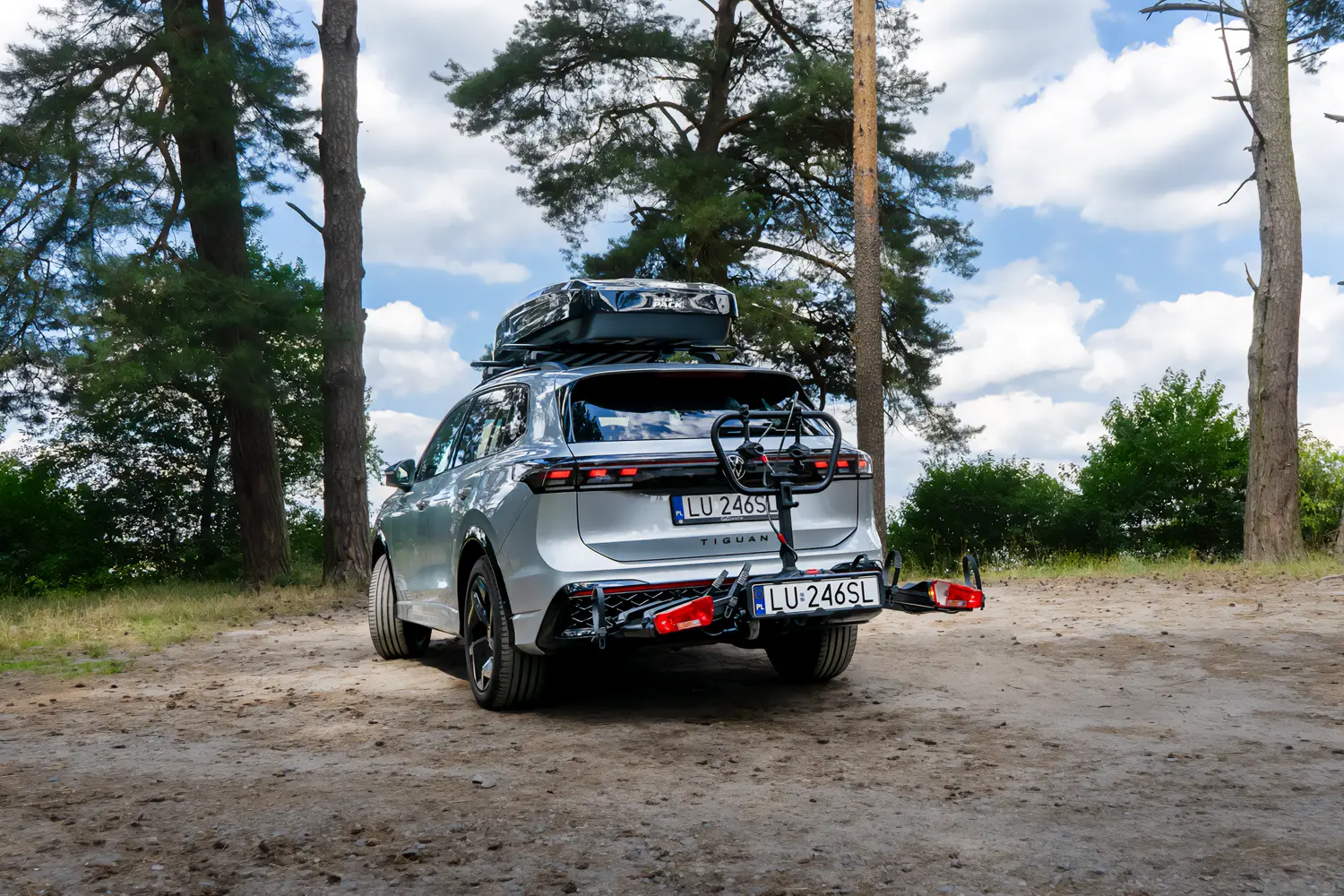 Silberner VW Tiguan mit einer Inter Pack Dachbox und einem Fahrradträger für die Anhängerkupplung auf einem Waldweg.
