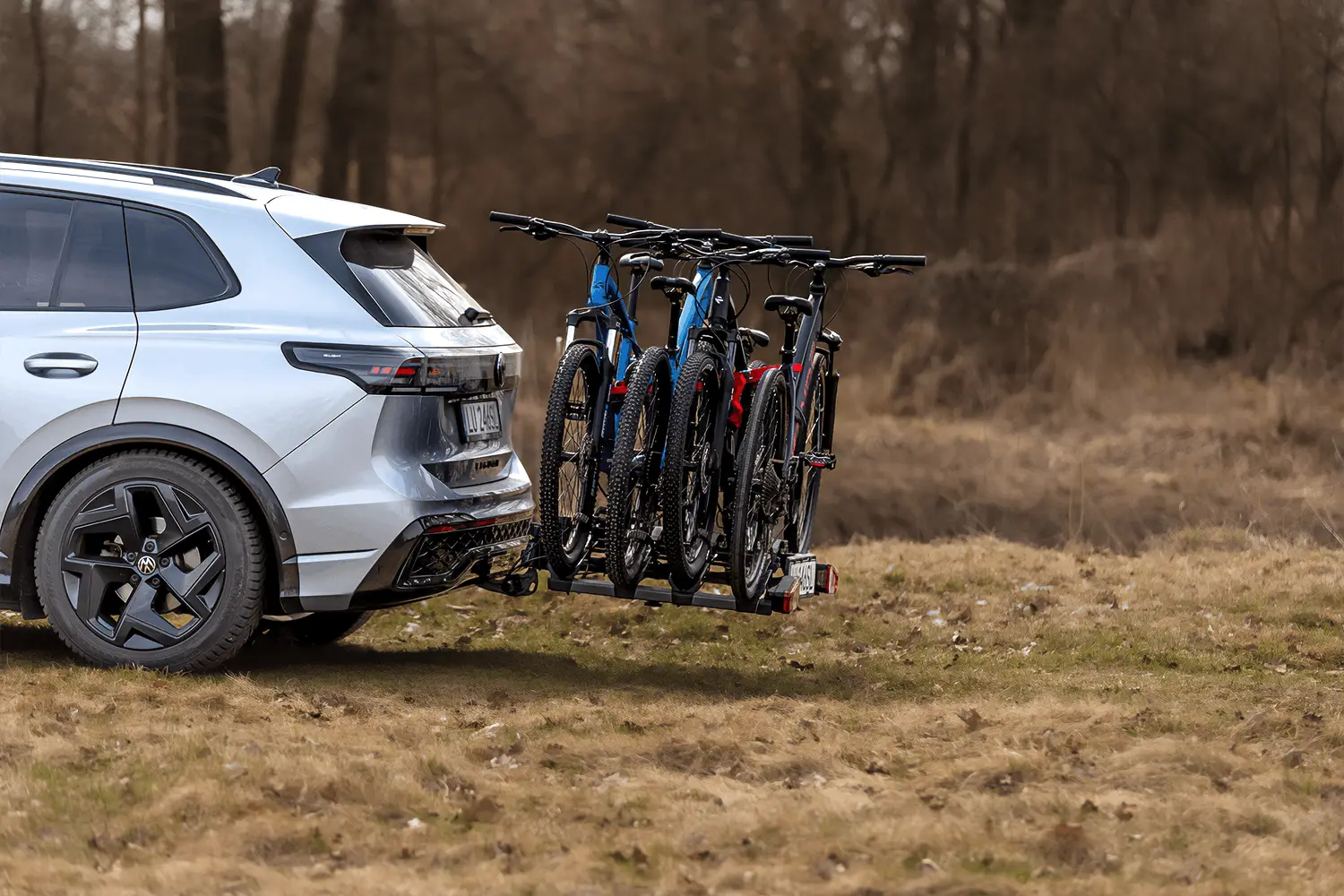 Silberner SUV mit einem beladenen Kupplungs-Fahrradträger für vier Mountainbikes auf einer Grasfläche am Waldrand.