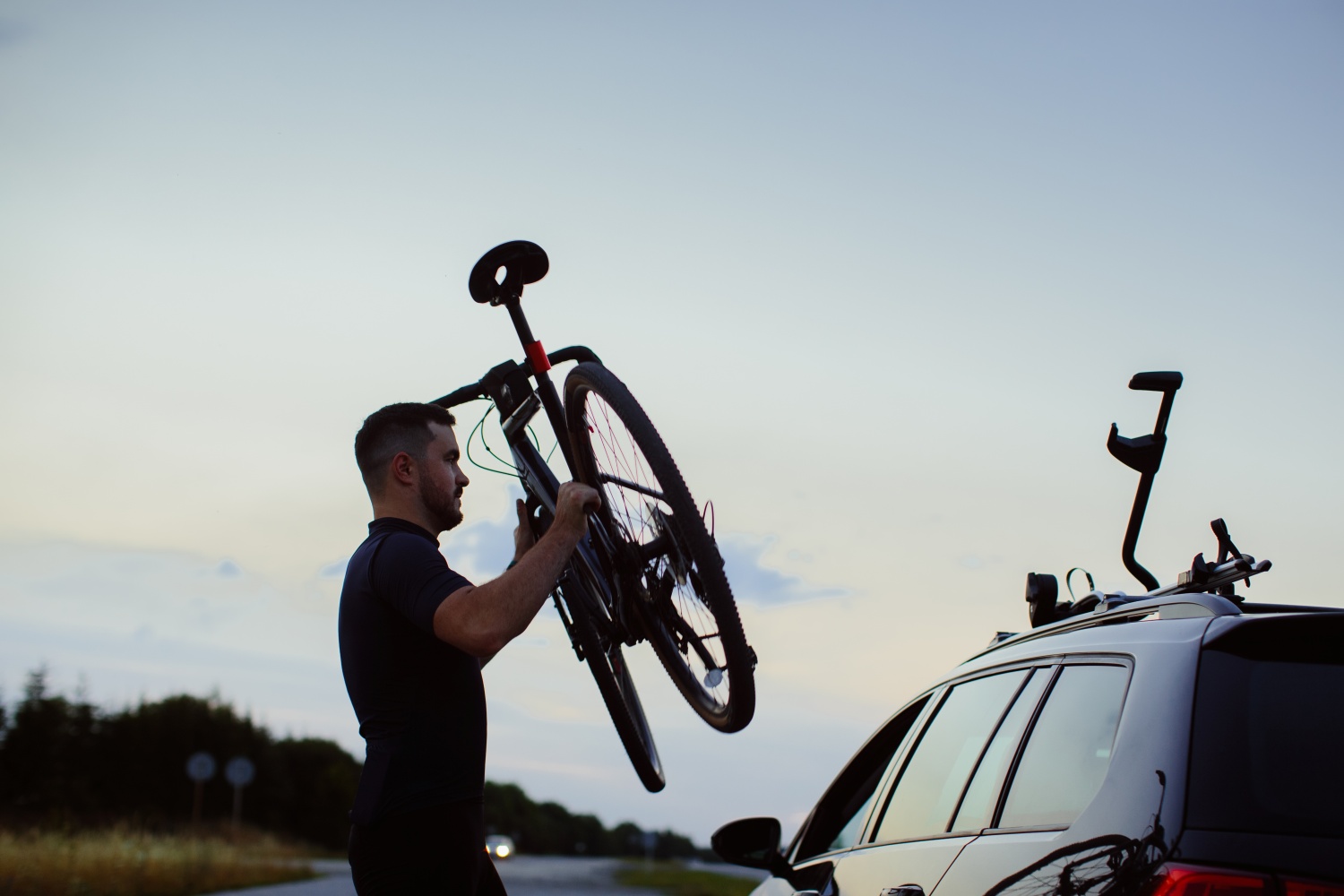 Ein Mann hebt im Abendlicht ein Fahrrad an, um es auf dem Dachträger eines Autos zu montieren.